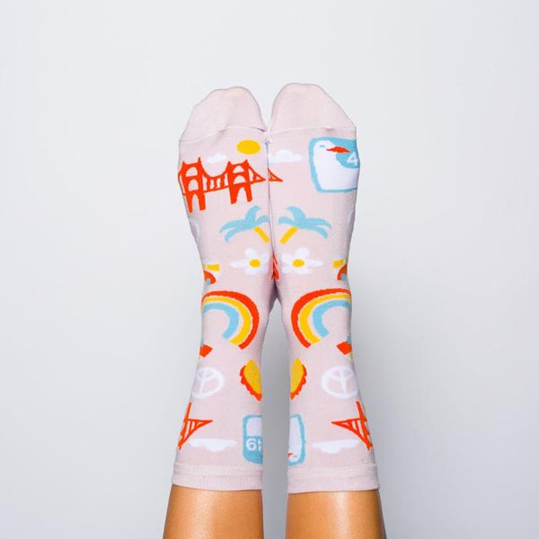 Shown on a models feet with a top down view, a pair of women's Yellow Owl Workshop cotton and nylon crew socks in lilac. These socks feature iconic San Francisco imagery like the golden gate bridge, peace signs, VW vans, seagull signage, and flower power.