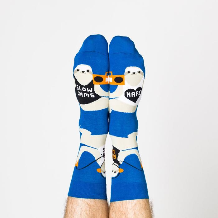 Model wearing a pair of Yellow Owl Workshop’s royal blue nylon-cotton men's crew socks with large sloths listening to music, holding boomboxes and wearing shirts that say “Sloth Life,” “Naps,” “Slow Jams” or “Weekends”